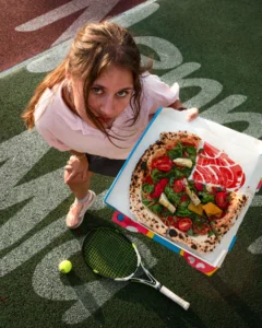 Pizza napolitaine fumante sur table, écran de foot en arrière-plan, verres levés